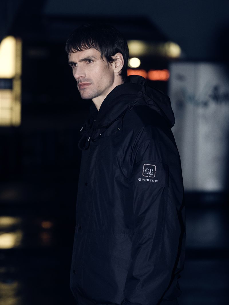Portrait of a man in a dark hooded jacket, looking off-camera in an urban setting with blurred background lights at night.