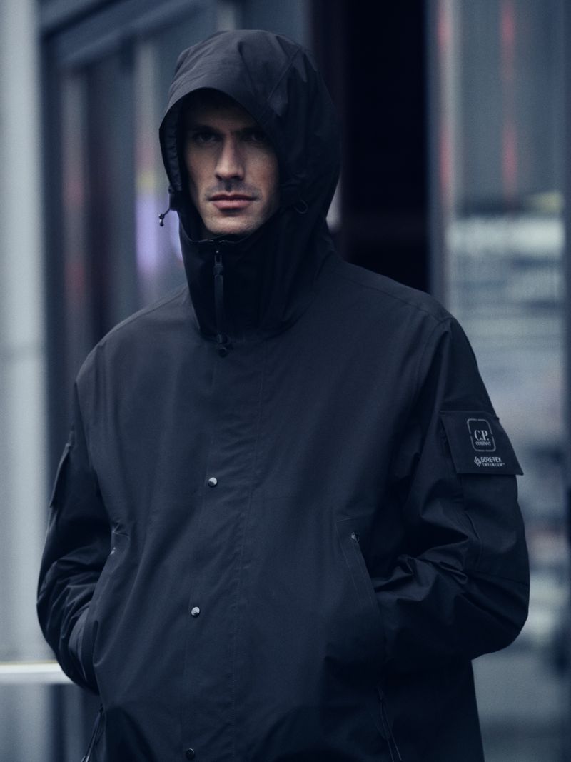 Male model in a black hooded jacket, posing in an urban setting, showcasing fashion in a portrait shot, featuring C.P. Company logo.