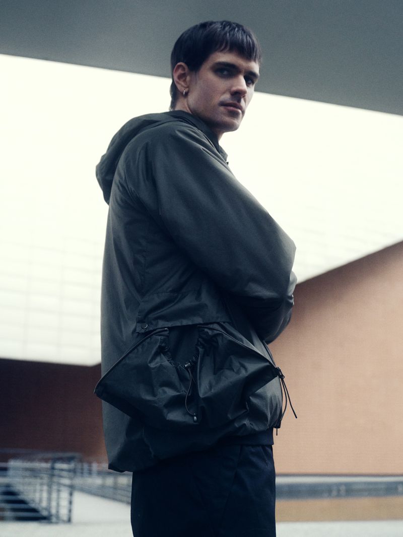 A male model poses outdoors wearing an olive-green hooded jacket and dark pants, glancing over his shoulder towards the camera.