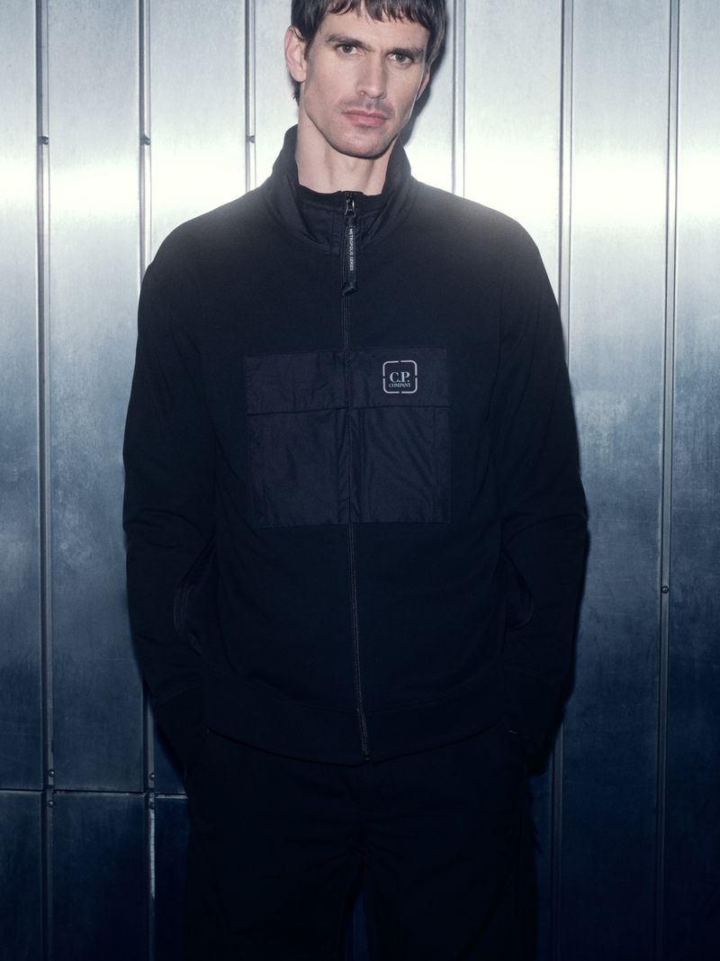 A man in a black jacket with a logo on the front stands against a textured metal background indoors, showcasing fashion and style.