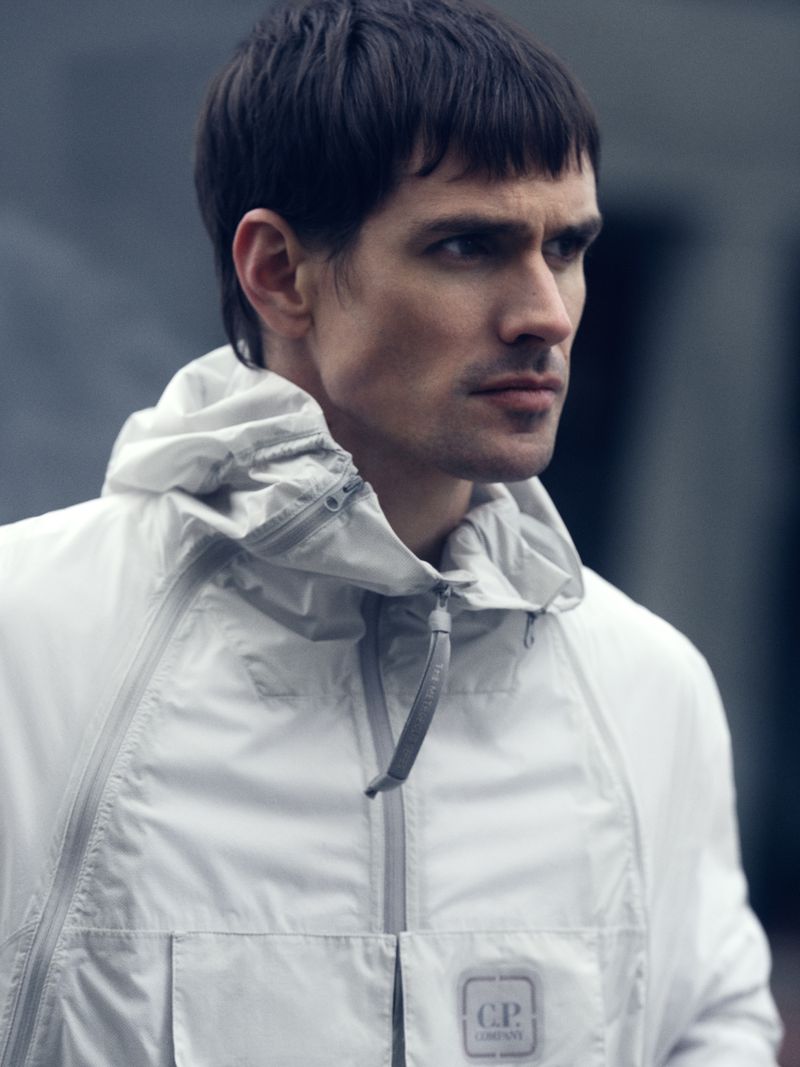 Close-up studio portrait of a handsome adult male in a white hooded jacket with a logo on the pocket, medium shot, neutral expression.