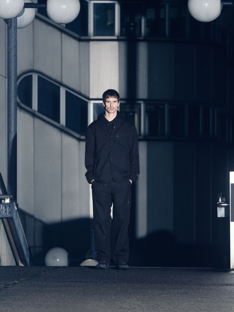 Adult man in dark clothing stands in an urban area at night, lit by globe lights near a building with a concrete and window facade.