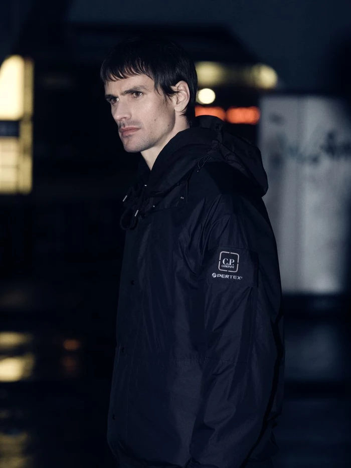 Man wearing a black hooded jacket with brand logo on sleeve against a dark urban street background at night