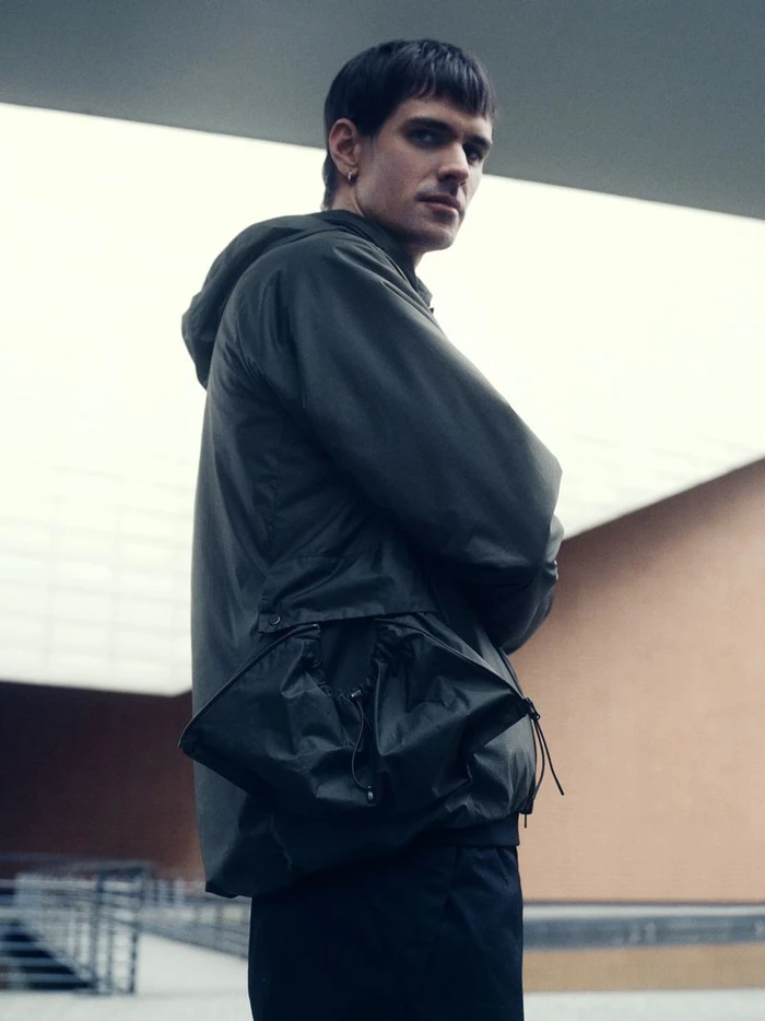 A man in a dark green hooded jacket with large front pockets standing against an architectural background.