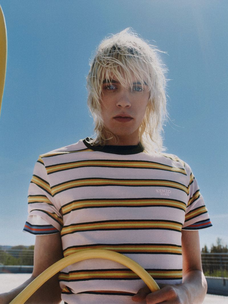 Portrait of a young adult with blonde hair wearing a striped t-shirt, holding a yellow tube, set against a bright blue sky outdoors.
