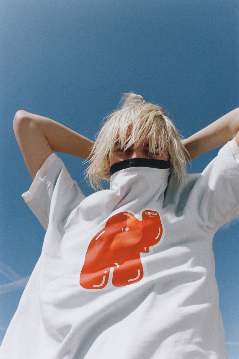 Fashion shot of a blonde adult woman in a graphic tee with hands behind her head, posing against a clear blue sky backdrop.