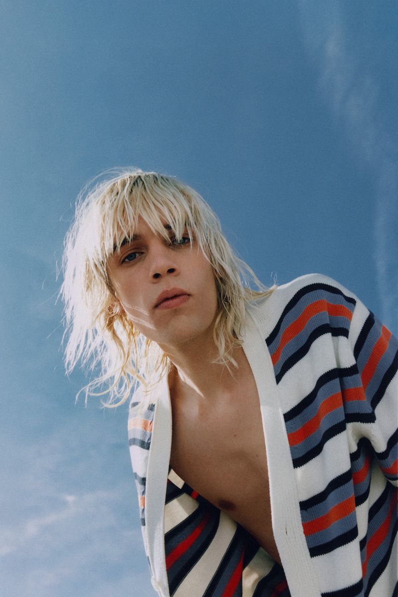 Low-angle shot of a young adult with blonde hair wearing a striped cardigan against a clear blue sky, creating a modern fashion portrait.