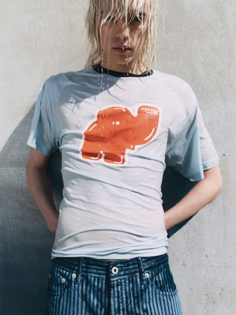 Androgynous model with wet blond hair wearing a wet Kenzo graphic tee and jeans in front of a concrete background, hands on hips.