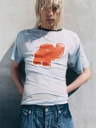 Androgynous model with wet blond hair wearing a wet Kenzo graphic tee and jeans in front of a concrete background, hands on hips.