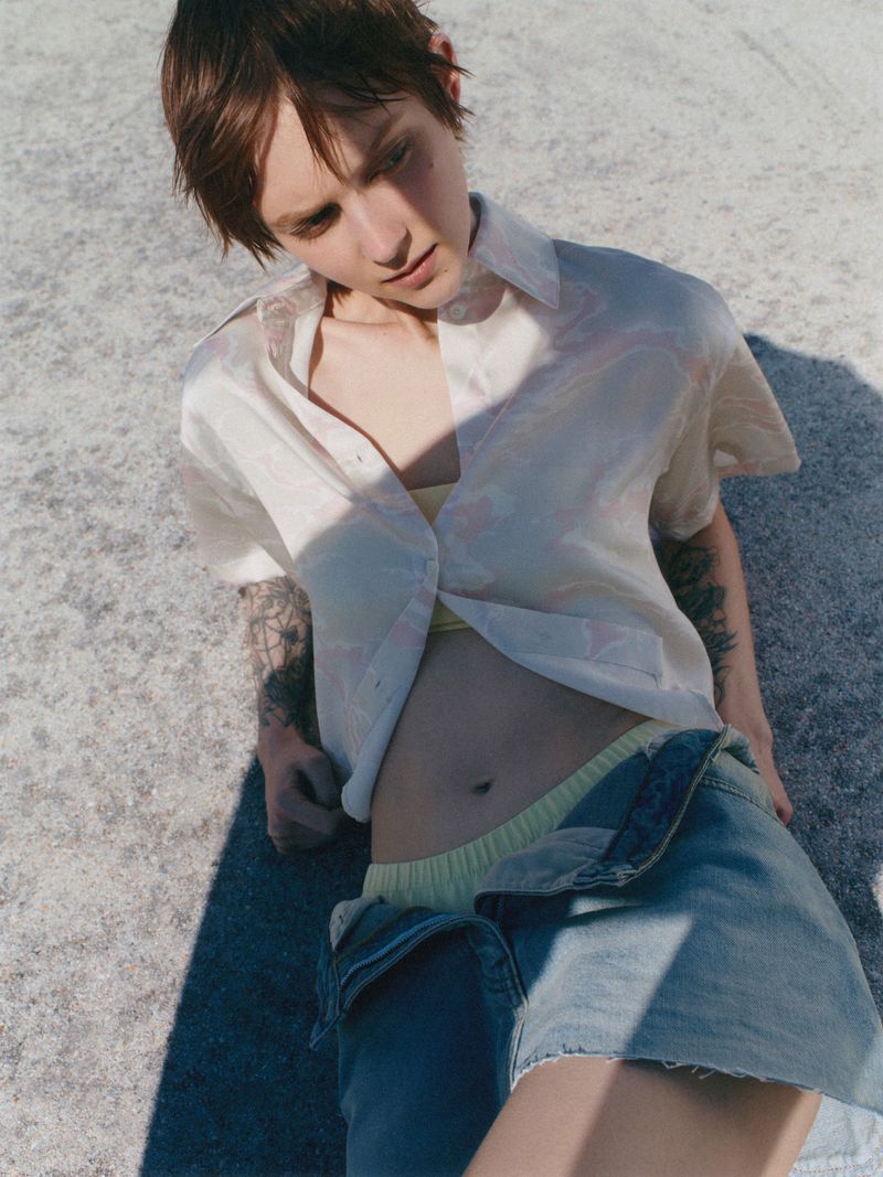 A young adult woman lying on the ground wearing a short denim skirt and an unbuttoned patterned shirt in a sunny outdoor setting.