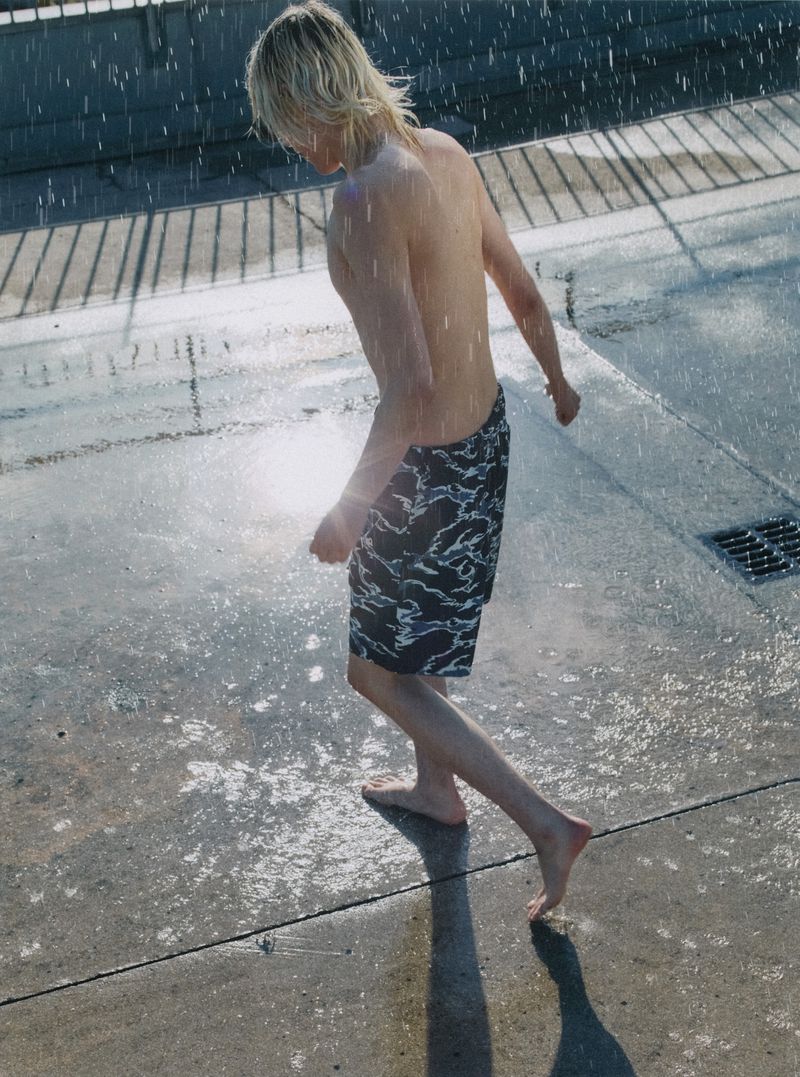 Blonde male barefoot and bare-chested walks in rain, wearing patterned shorts on a concrete surface with a grated drain.