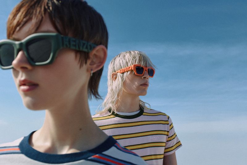 Two people wear 'KENZO' logo sunglasses. The first has dark hair, the second with blonde mullet and striped shirt, clear sky background.