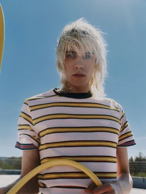 Model with blonde hair wearing a striped t-shirt holding a yellow object against a clear blue sky background