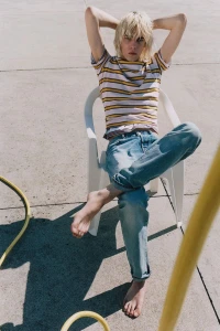 Model with blonde hair seated on a white plastic chair wearing a striped t-shirt and blue jeans