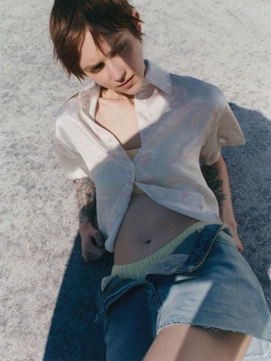 Model with short brown hair wearing a light patterned button-down shirt and denim skirt against a concrete background.