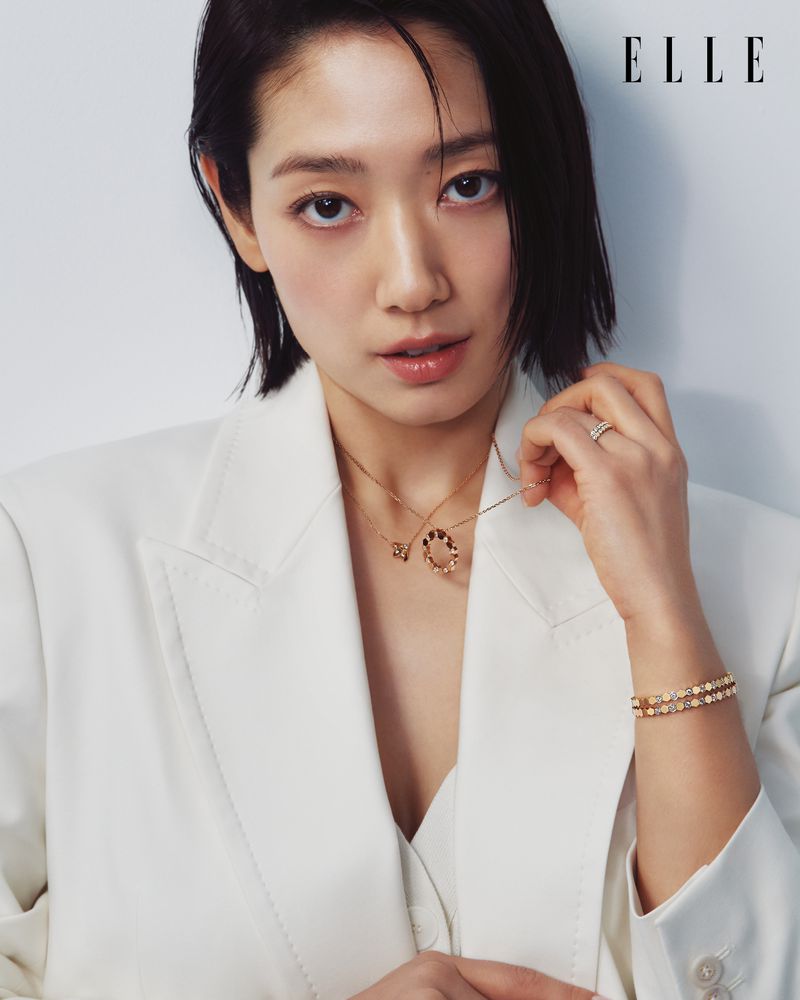Portrait of a young Asian woman with short dark hair wearing a white blazer with two necklaces, bracelet, and ring against a white backdrop.