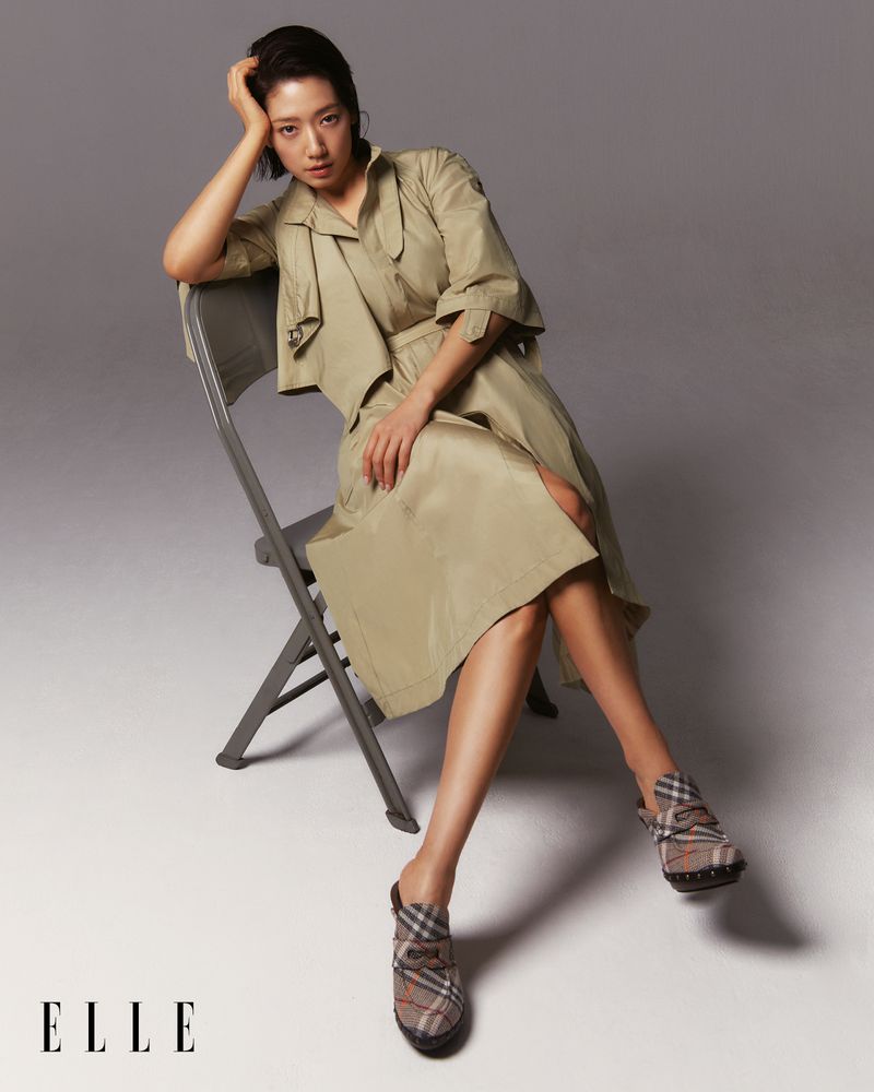 An Asian woman with short brown hair is seated on a grey folding chair, wearing a beige dress and patterned brown heeled shoes.