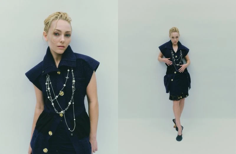 A woman in a short, dark blue dress with a layered necklace poses in front of a pale background in a fashion editorial shot.