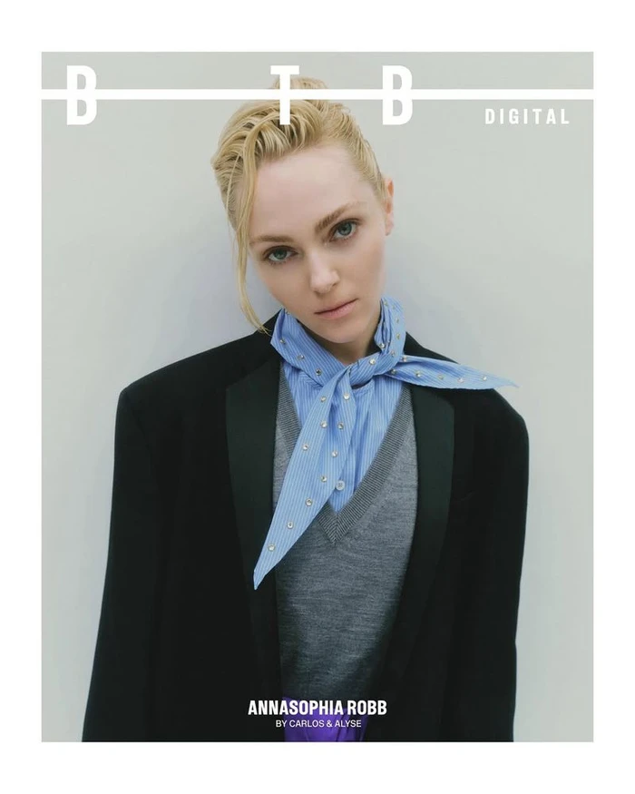 Portrait of Annasophia Robb wearing a black blazer, grey vest, striped blue neck scarf against a neutral wall