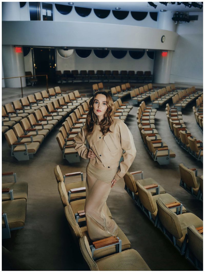 A young woman poses in a beige jacket and skirt in an auditorium with rows of empty seats, looking directly at the camera.