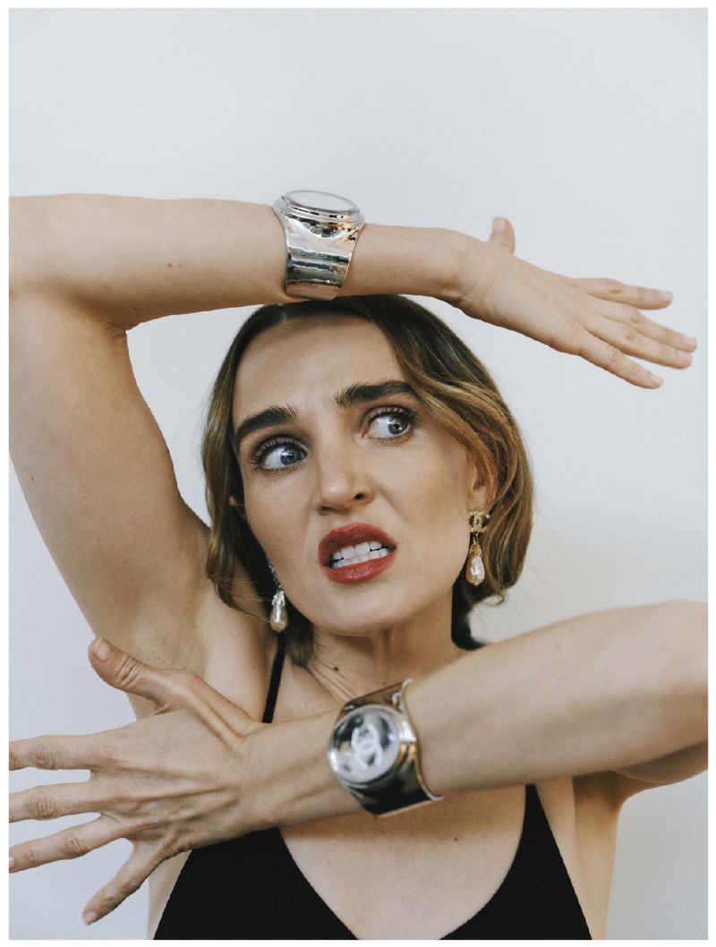 Woman with bright red lipstick poses indoors wearing two large watches on both wrists and dangle earrings, against a white wall.