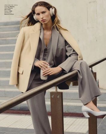 Adult woman in beige blazer and grey suit with headphones poses on stairs with railing holding cup in an urban environment.