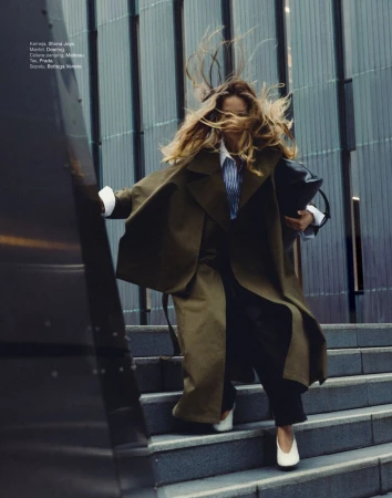 Woman in olive overcoat walks down steps outside a building. She holds a black bag. Her hair is windswept. She wears white flats.