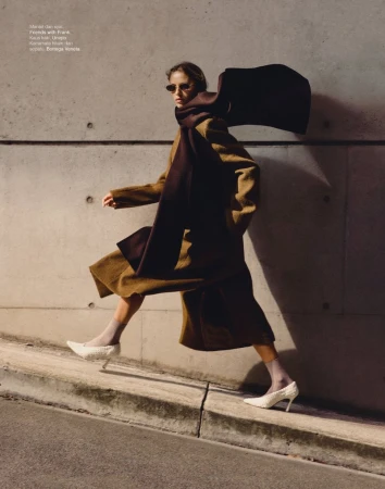 Fashion editorial shot of a woman walking outdoors wearing a brown coat, brown scarf, and white heels, posing against a concrete wall.