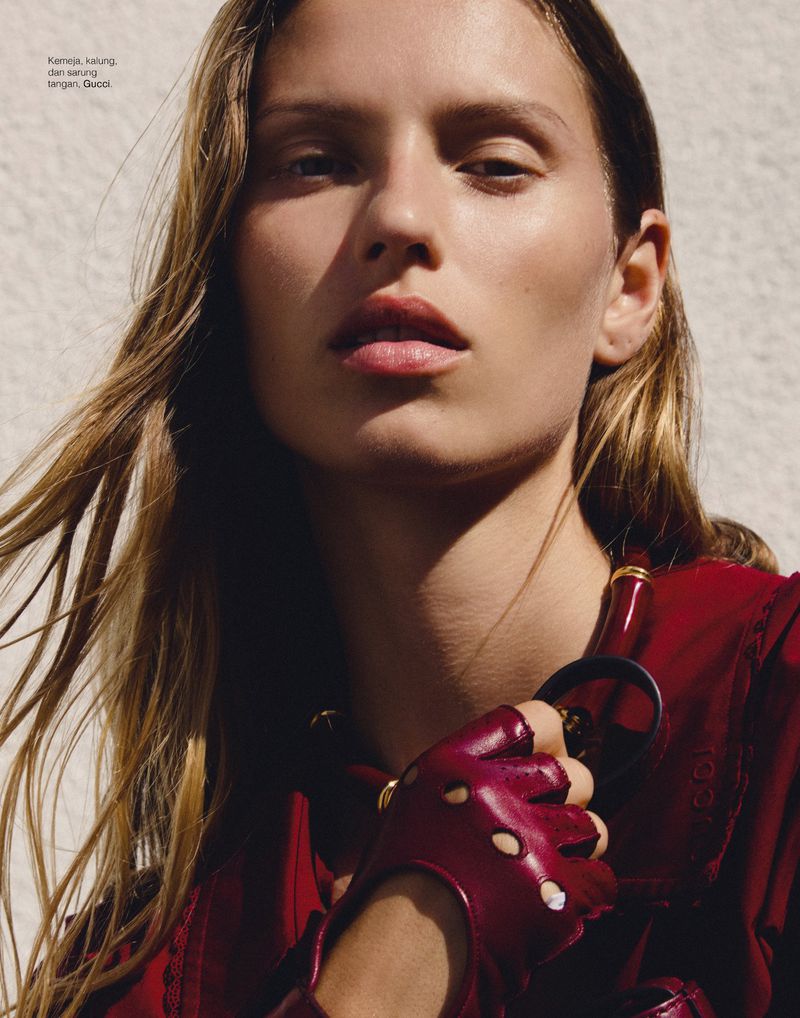 Close-up fashion portrait of a fair-skinned adult woman wearing a red top, leather gloves, and gold jewellery against a neutral background.