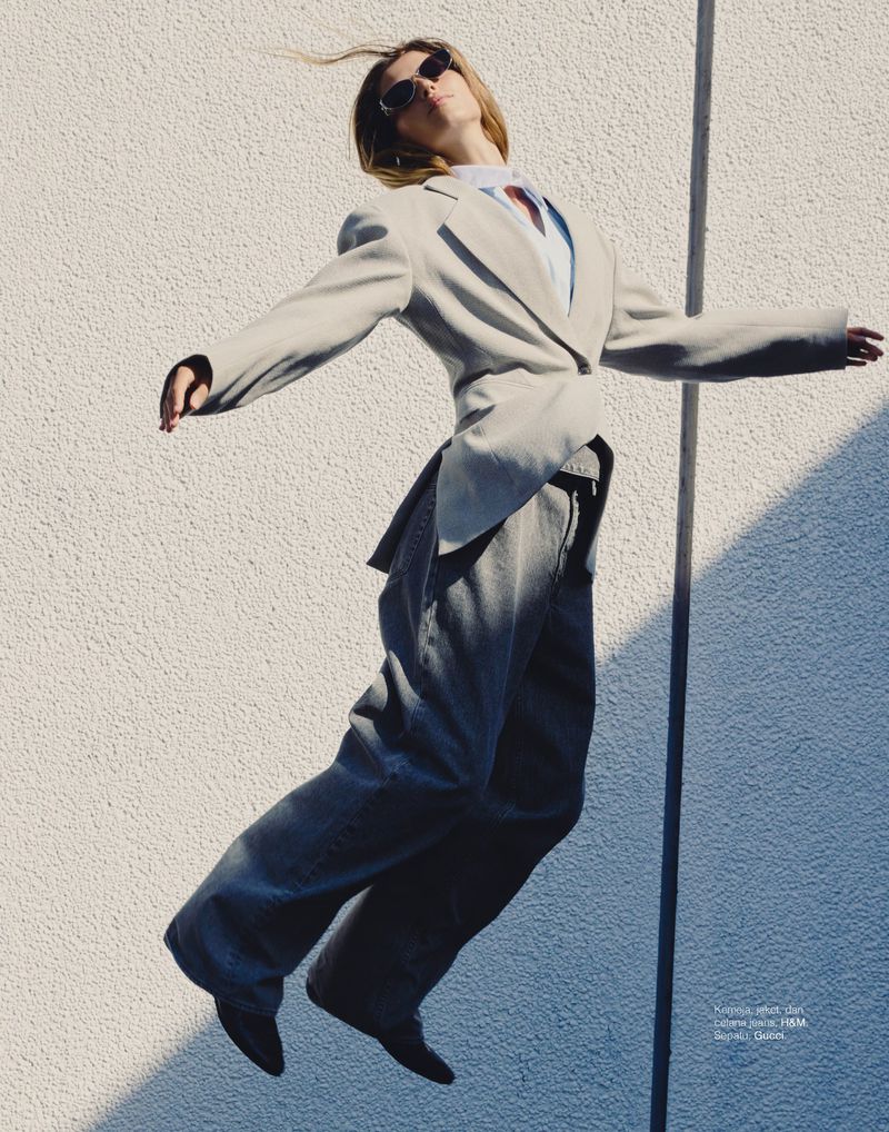 Woman in a blazer and jeans jumps in front of a textured wall, wearing sunglasses, capturing a moment of movement and fashion.