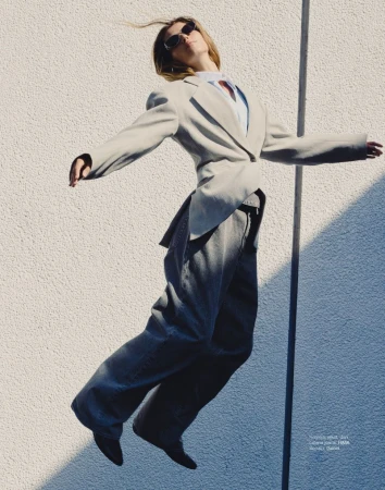 Woman in a blazer and jeans jumps in front of a textured wall, wearing sunglasses, capturing a moment of movement and fashion.