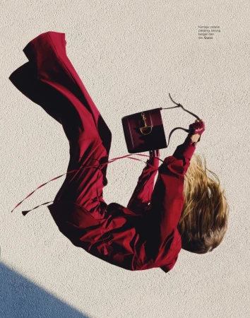 Fashionable woman in red jumpsuit levitates while holding a burgundy leather purse against a beige textured background, vertical.