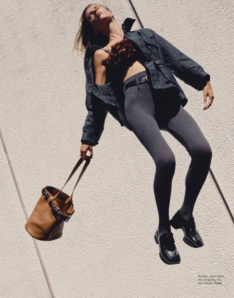Fashion shot of a woman wearing a jacket, top, and leggings, holding a bucket bag against a textured wall in an outdoor setting.