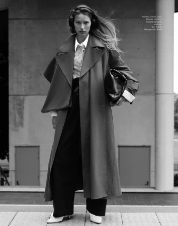 A woman in a trench coat and headphones stands against a concrete background, carrying a black purse in a fashion editorial.