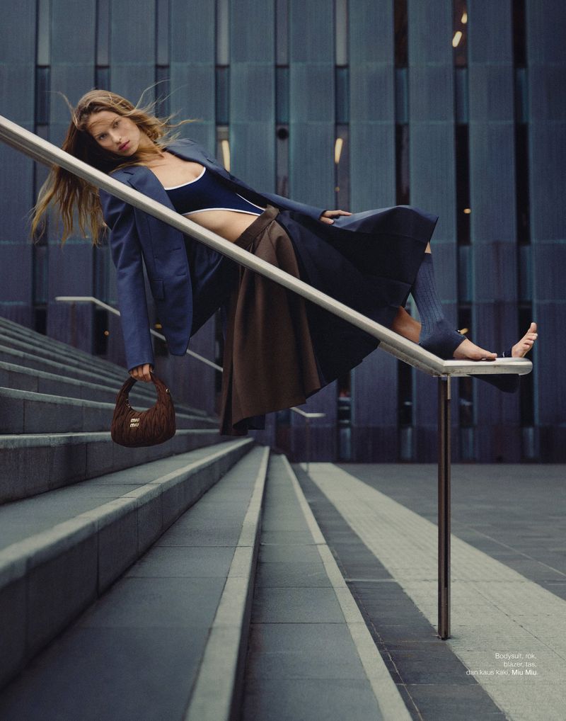 Fashion shot of a woman posing on stairs in a navy jacket, crop top, and brown skirt, holding a furry handbag in daylight.