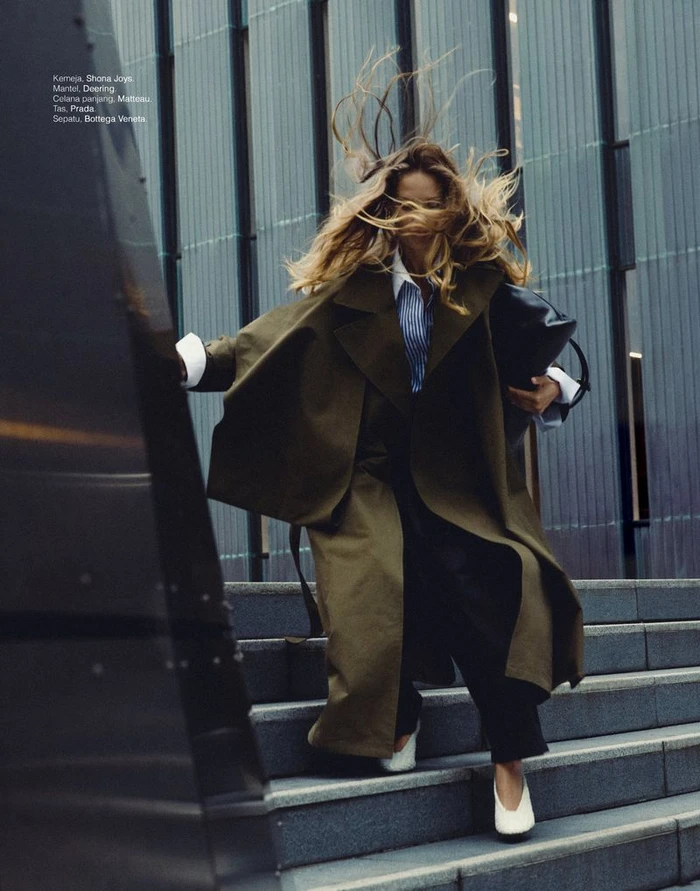 Model Lea Seelenmeyer descending outdoor stairs in a long olive coat with wind whipping her blonde hair, for Elle Indonesia.