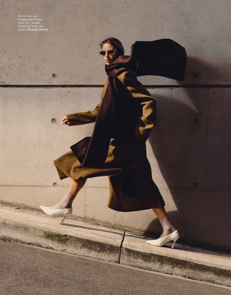 Model wearing a brown coat and dark sunglasses walking against a concrete wall for Elle Indonesia