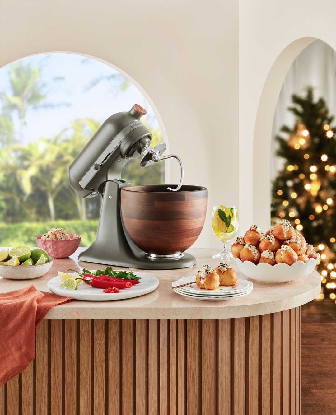 KitchenAid stand mixer with a wooden bowl on a countertop surrounded by limes, peppers, appetizers, and Christmas tree in background.