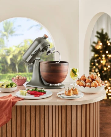 KitchenAid stand mixer with a wooden bowl on a countertop surrounded by limes, peppers, appetizers, and Christmas tree in background.