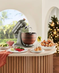 Grey stand mixer with wooden bowl on a curved kitchen counter with holiday treats and a Christmas tree in the background