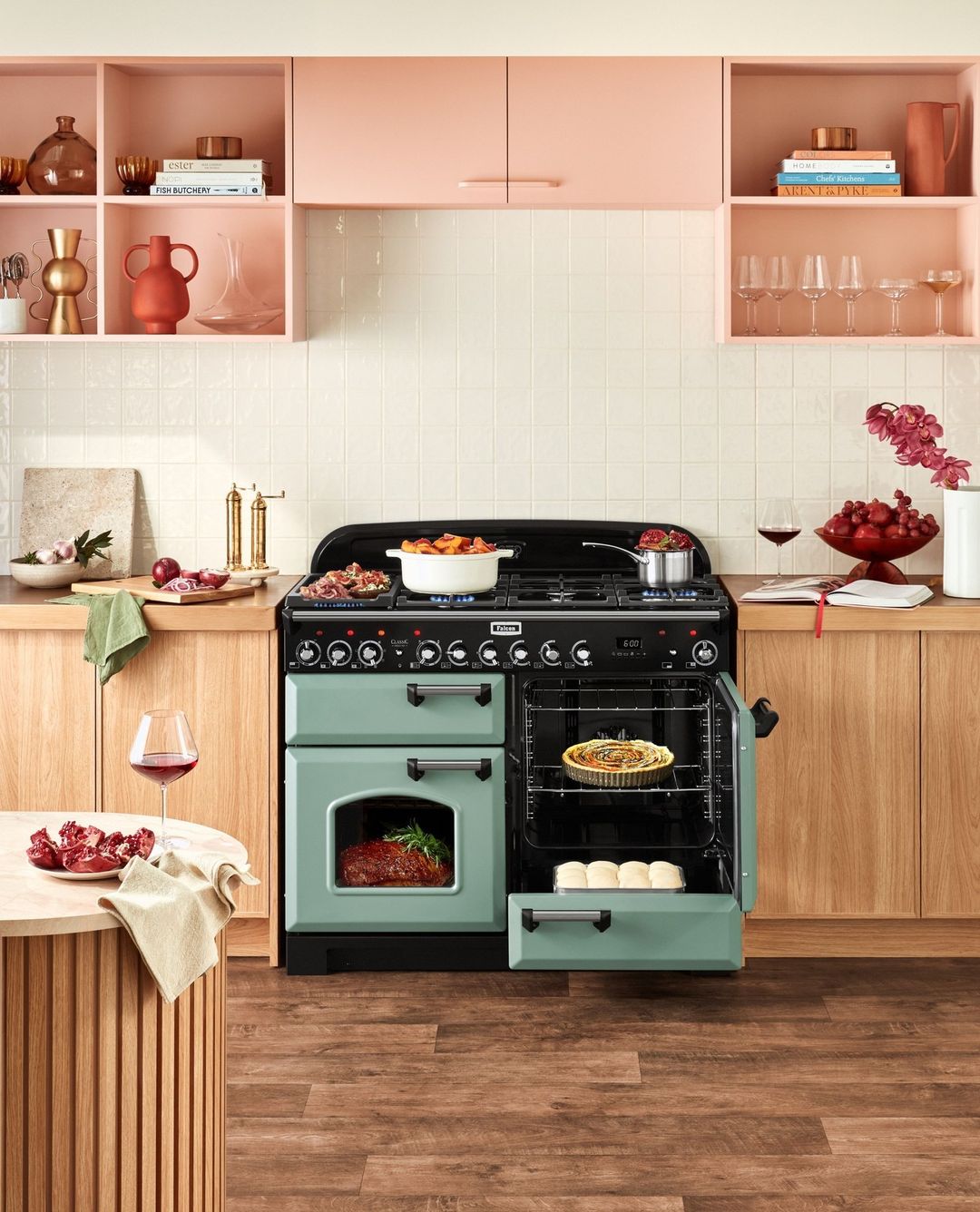 Kitchen interior with a green Falcon range oven and a variety of food items, including a pie, meat, vegetables, and bread rolls.