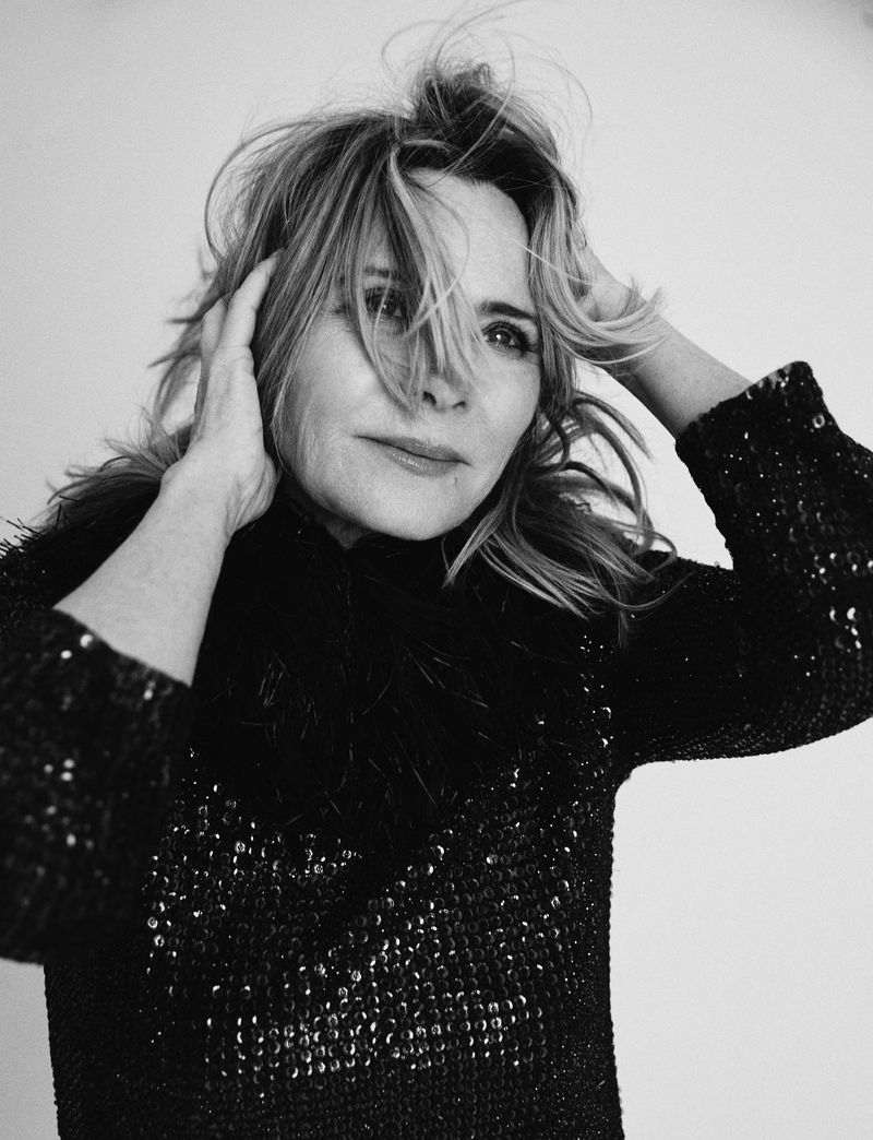 Black and white portrait of a mature woman with hands in her blond hair, wearing a sequined top with a feathered collar, against a white backdrop.