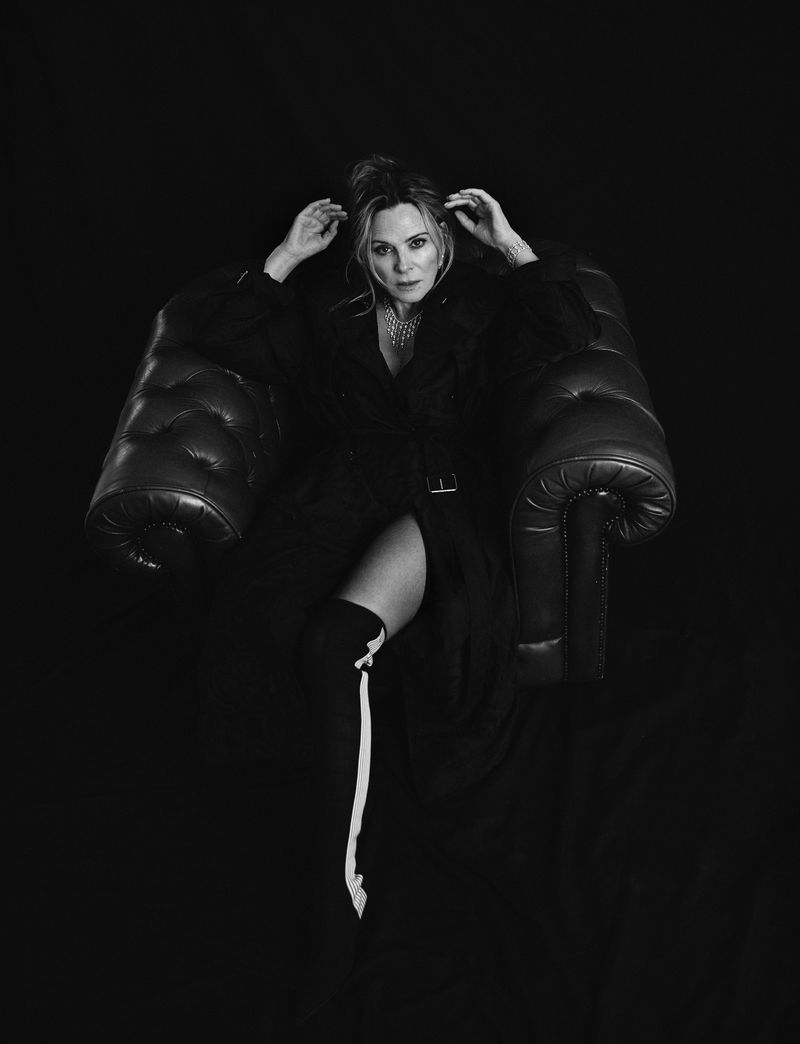 Black and white portrait of a woman in a long coat and thigh-high boots posing in a leather armchair against a dark background.