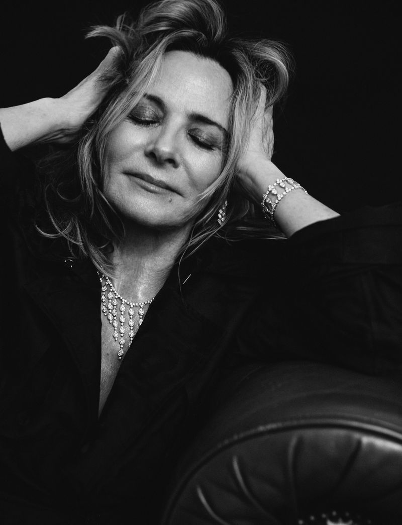 Black and white portrait of a mature adult woman with her hands in her hair, wearing a black jacket and diamond jewelry.