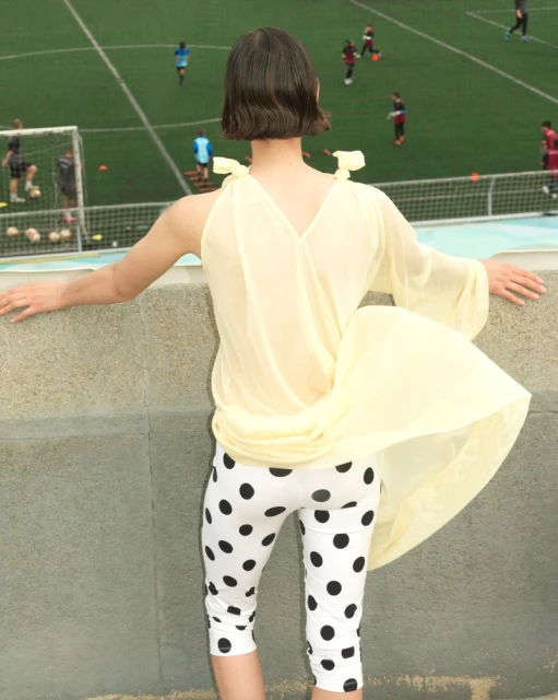 An adult poses outdoors wearing a sheer, pale yellow top, cropped polka dot pants with a sports field and players in the background.
