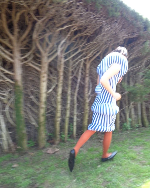 Woman in blue and white striped dress is running on green grass near bare trees, wearing red tights and black flats.