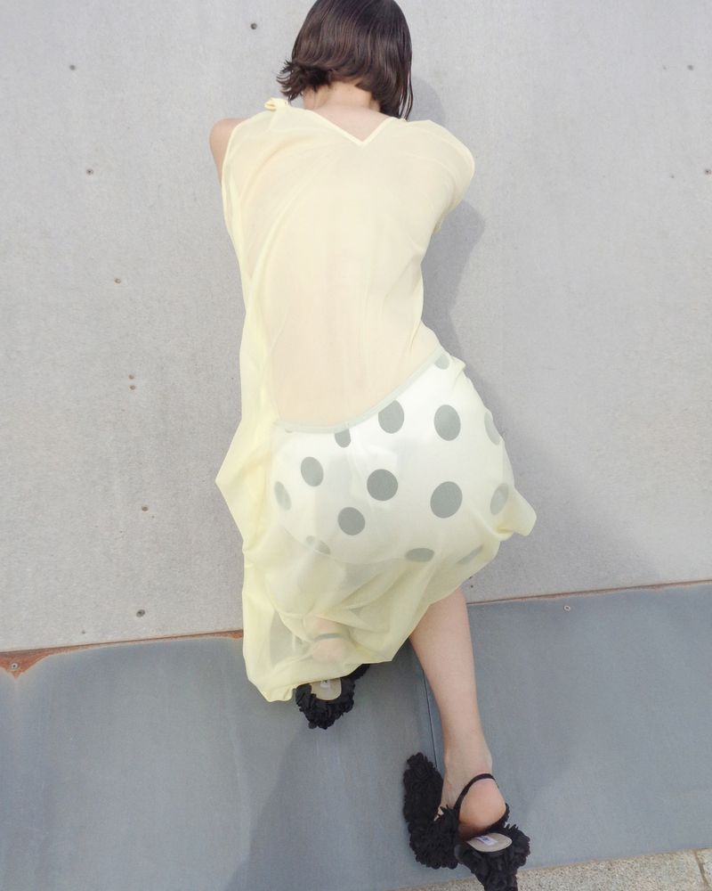 Back view of a woman in a pale yellow dress and black ruffled heels sitting against a gray concrete wall in soft, natural light.