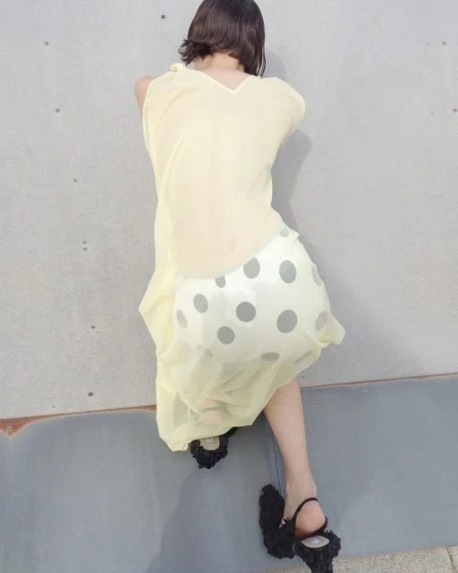 Back view of a woman in a pale yellow dress and black ruffled heels sitting against a gray concrete wall in soft, natural light.