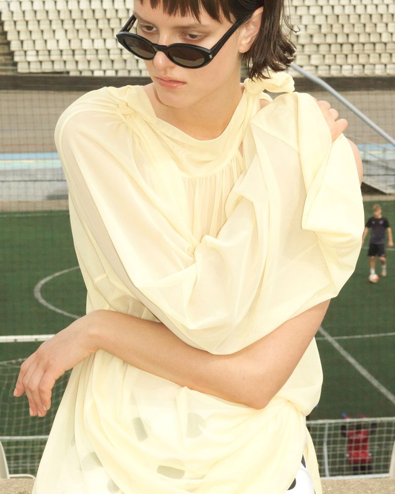 A young adult woman wears sunglasses and a light yellow dress, standing on a stadium concourse overlooking a green field.