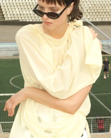 A young adult woman wears sunglasses and a light yellow dress, standing on a stadium concourse overlooking a green field.